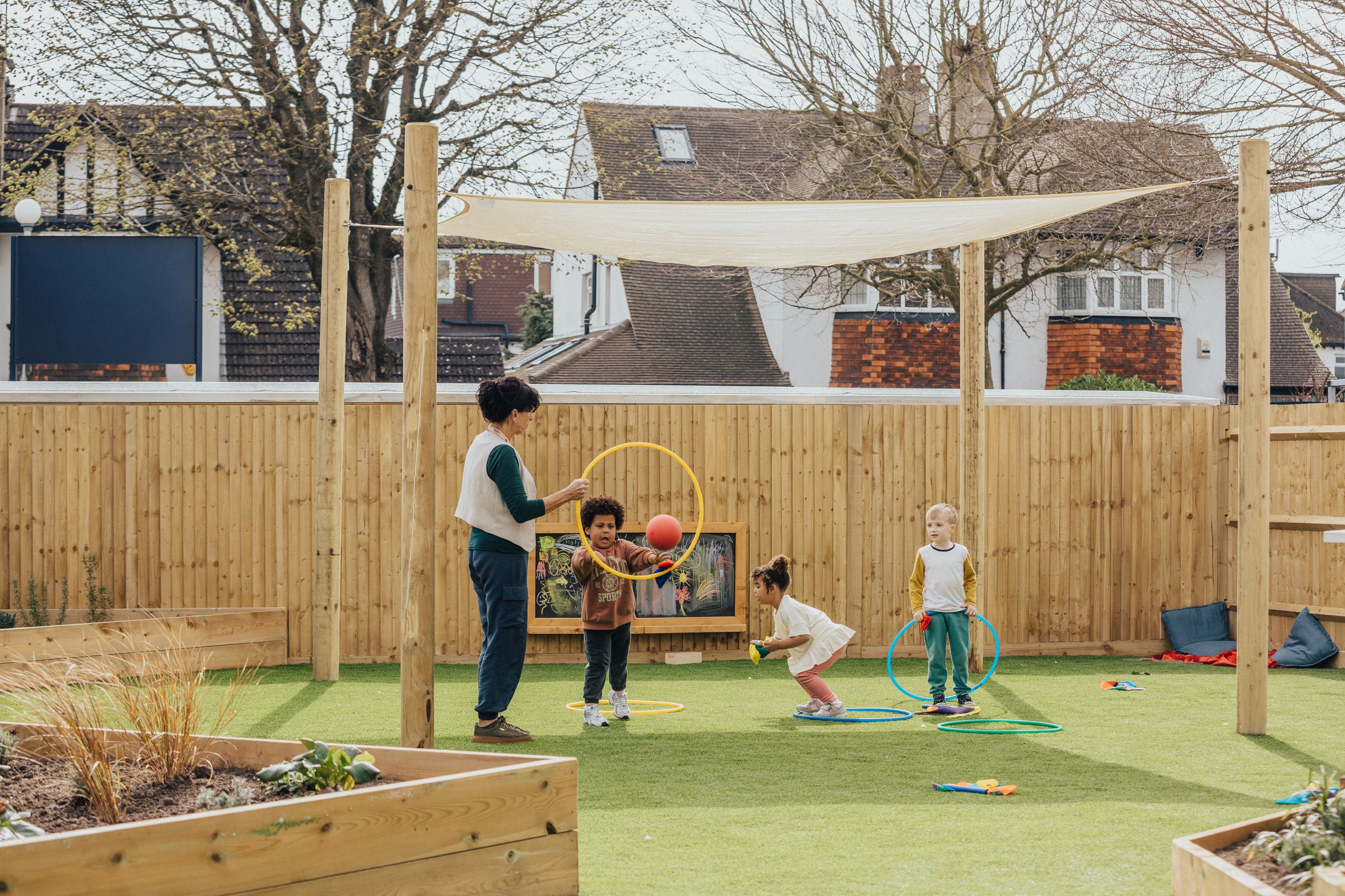 An adult and three young children play with hoops and balls on green turf in an outdoor playground with a wooden fence and shade sail.