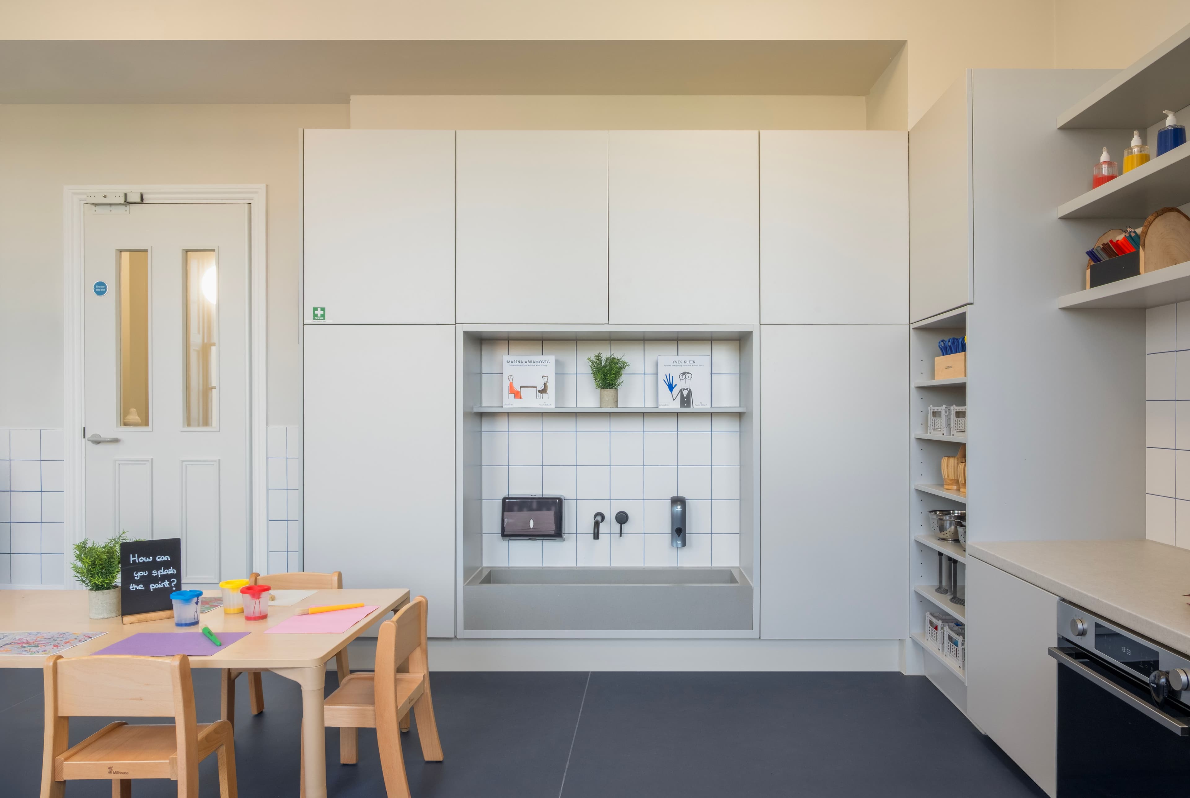 A modern children's room with a craft table, light grey cabinets featuring an integrated trough sink, and open shelves stocked with supplies.