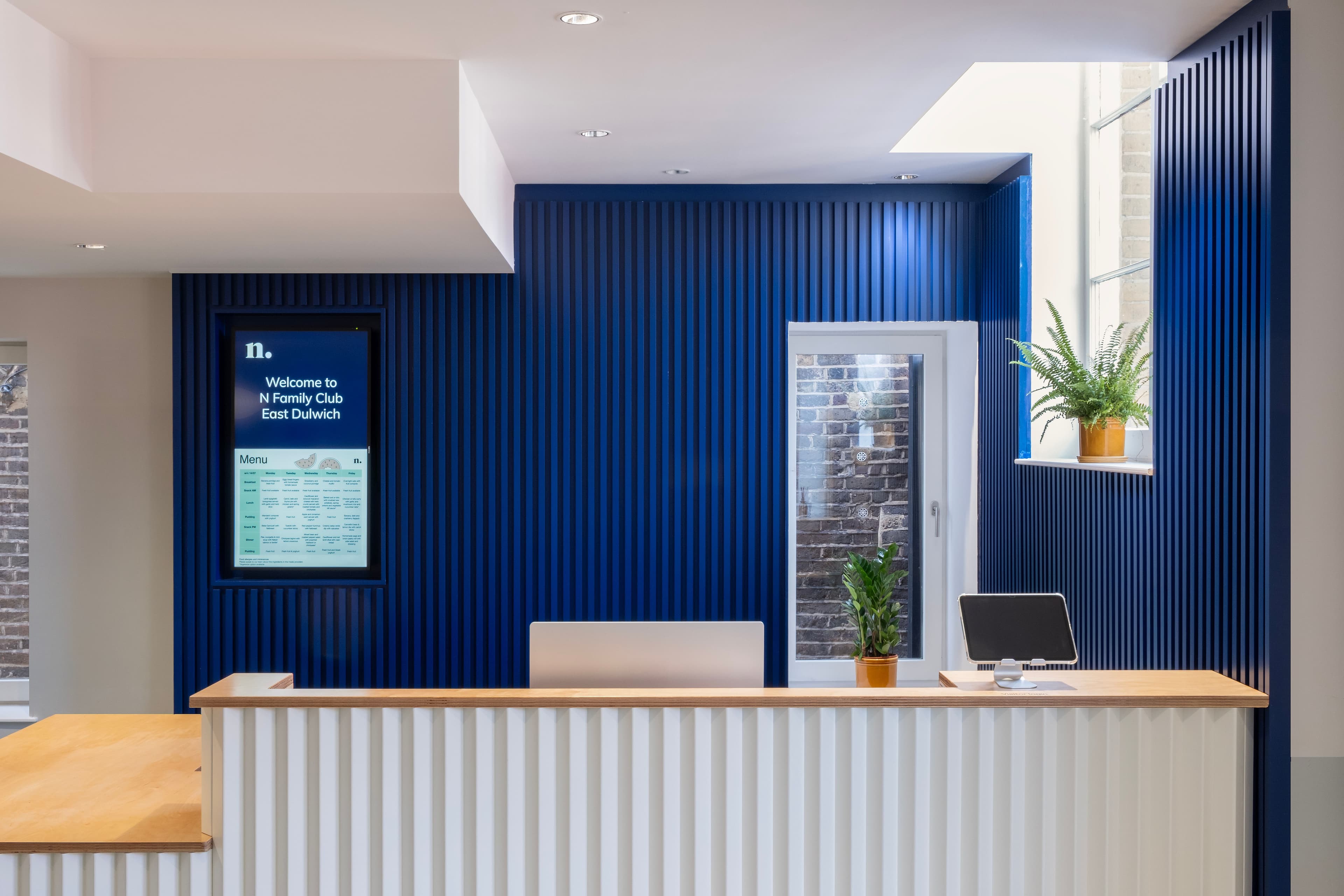 A modern reception area with a white and light wood desk, a dark blue slatted wall with a digital screen, and potted plants.