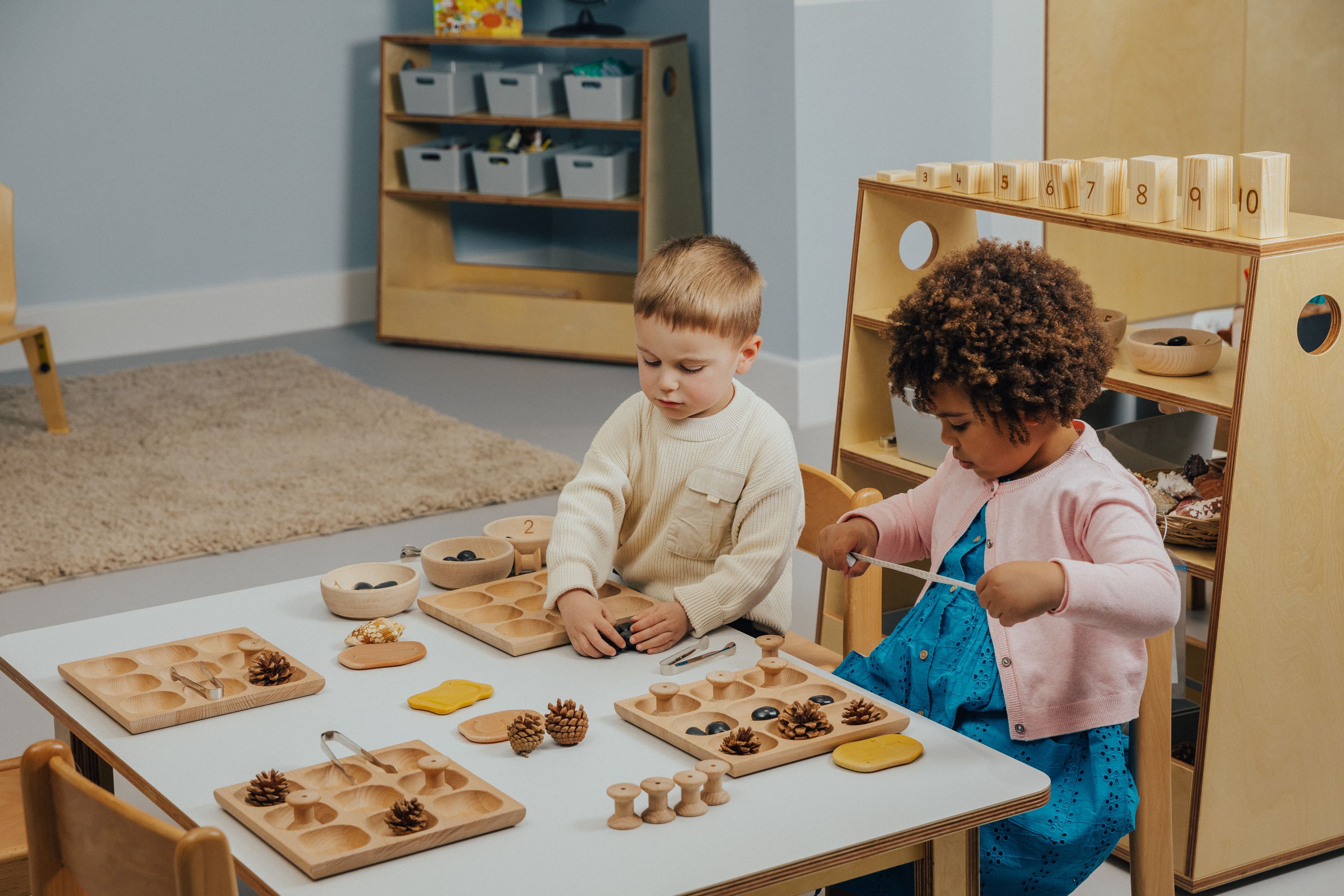 Two young children sort natural materials like pinecones and stones using wooden trays at a table in a classroom.