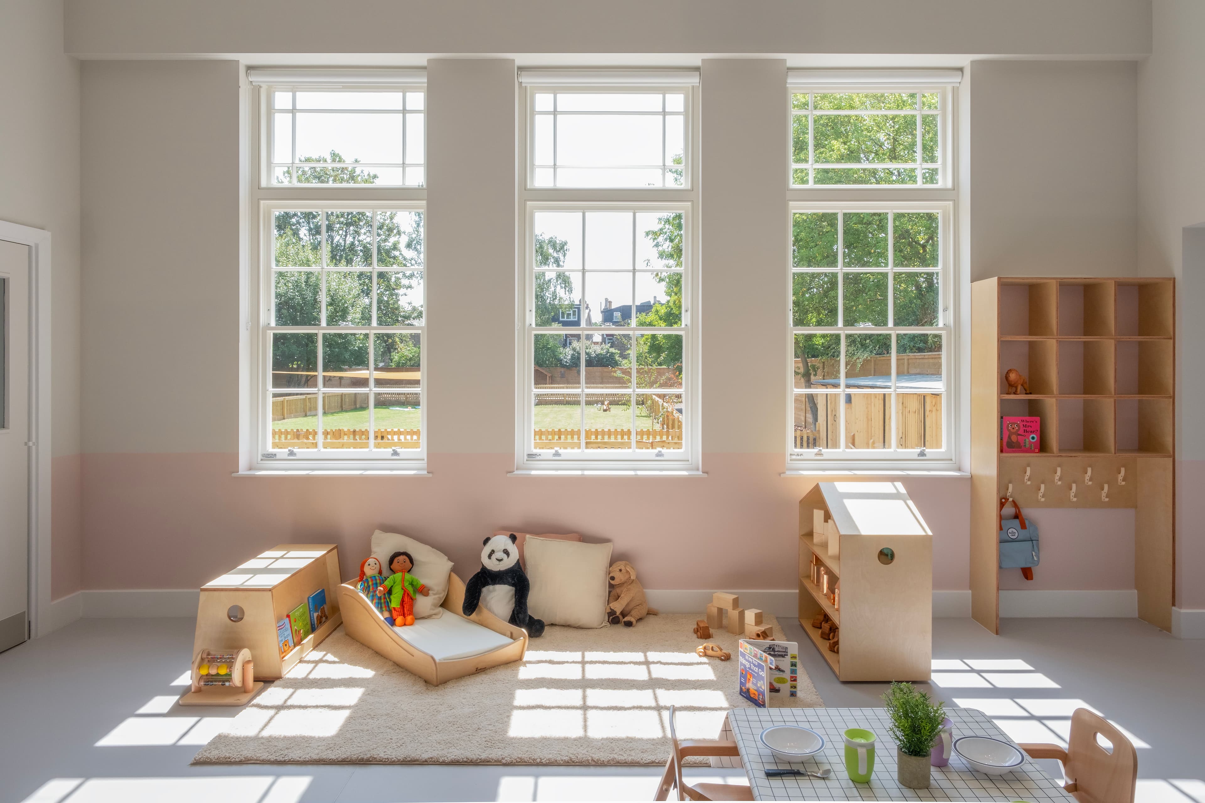 A bright, modern children's playroom with large windows, two-tone pink and beige walls, light wood furniture, and toys on a rug.