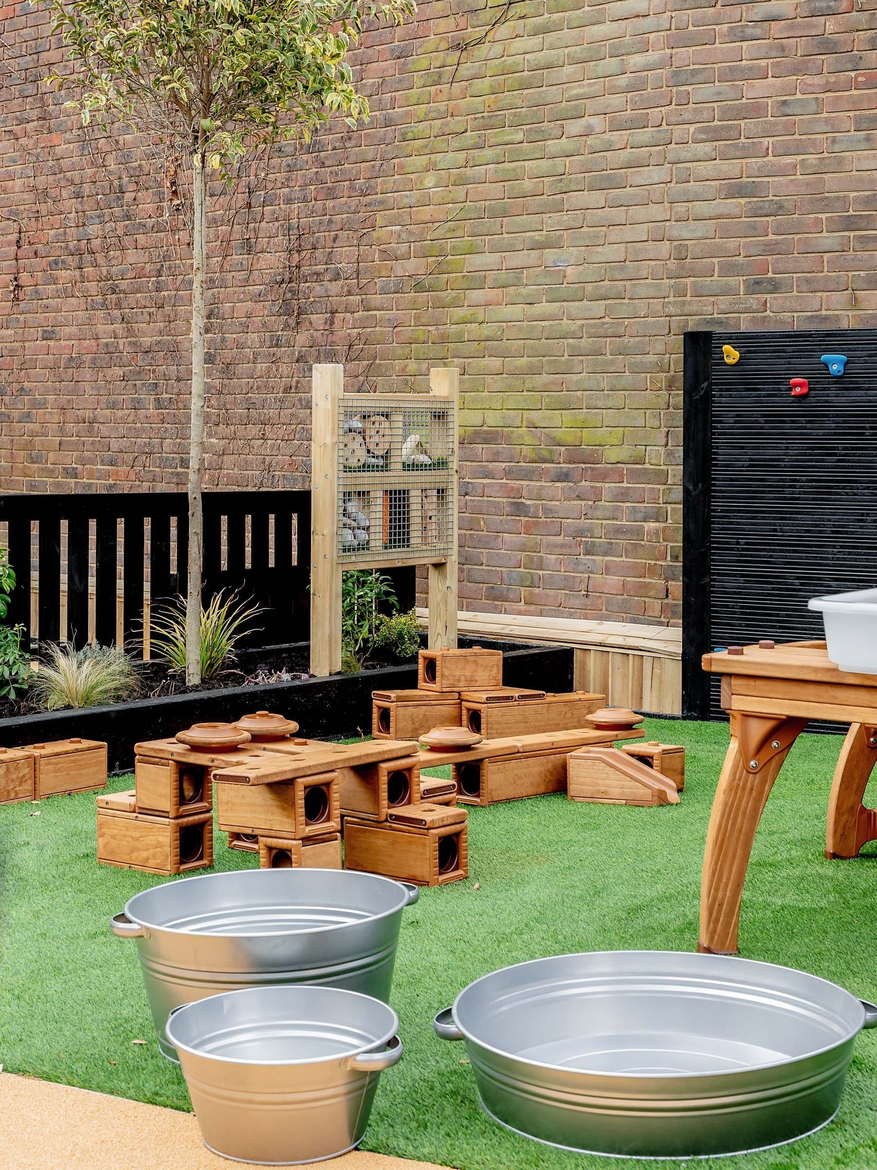 Outdoor play area with artificial grass, wooden block structures, metal tubs, an insect hotel, and a climbing wall against a brick backdrop.
