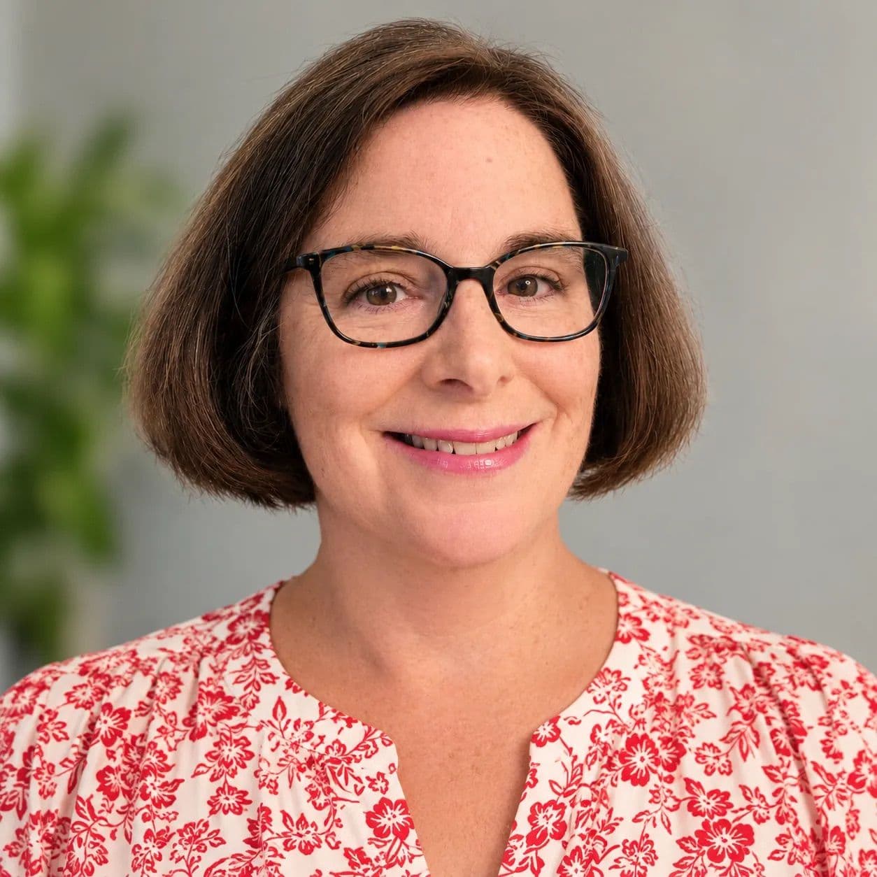 A smiling woman with short brown hair and tortoiseshell glasses wears a red and white floral top.