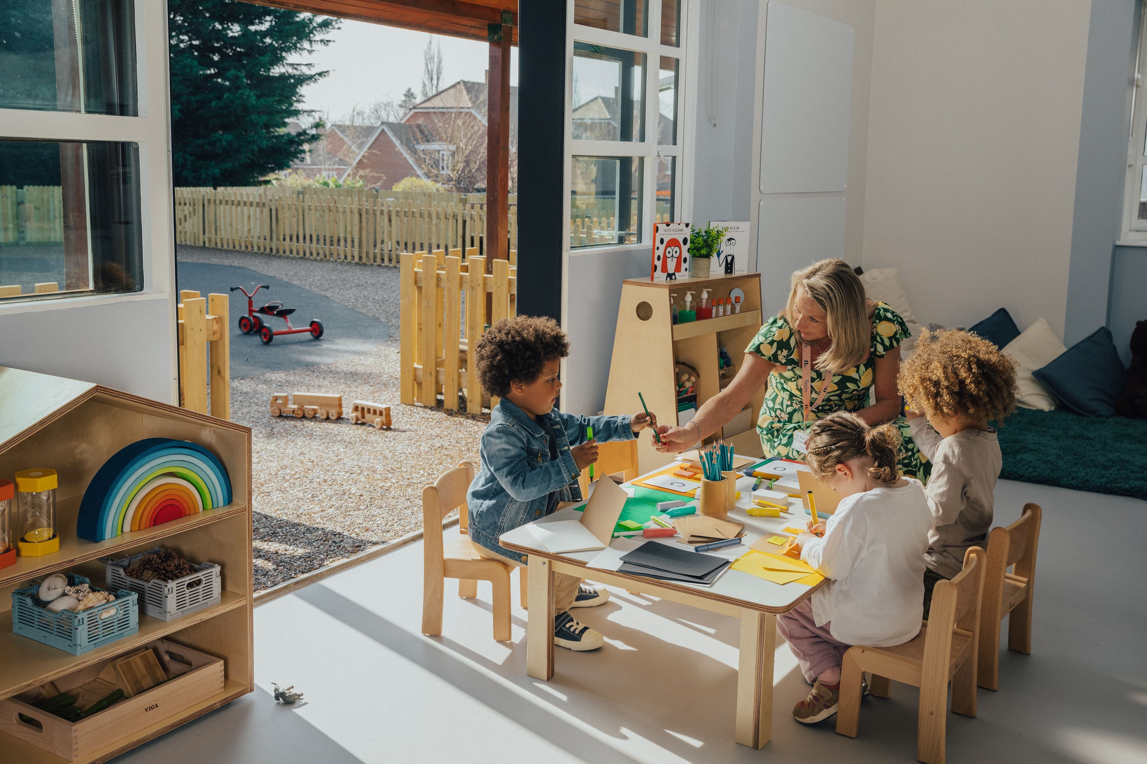 A teacher and three diverse children doing crafts at a table inside, with an outdoor play area visible.