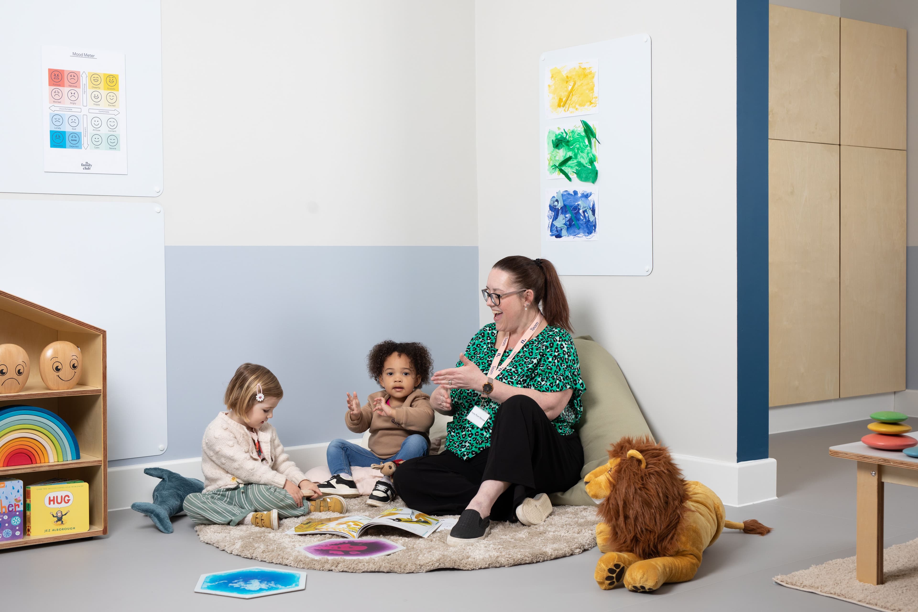 An adult woman claps while sitting on a rug with two toddlers in a bright classroom.