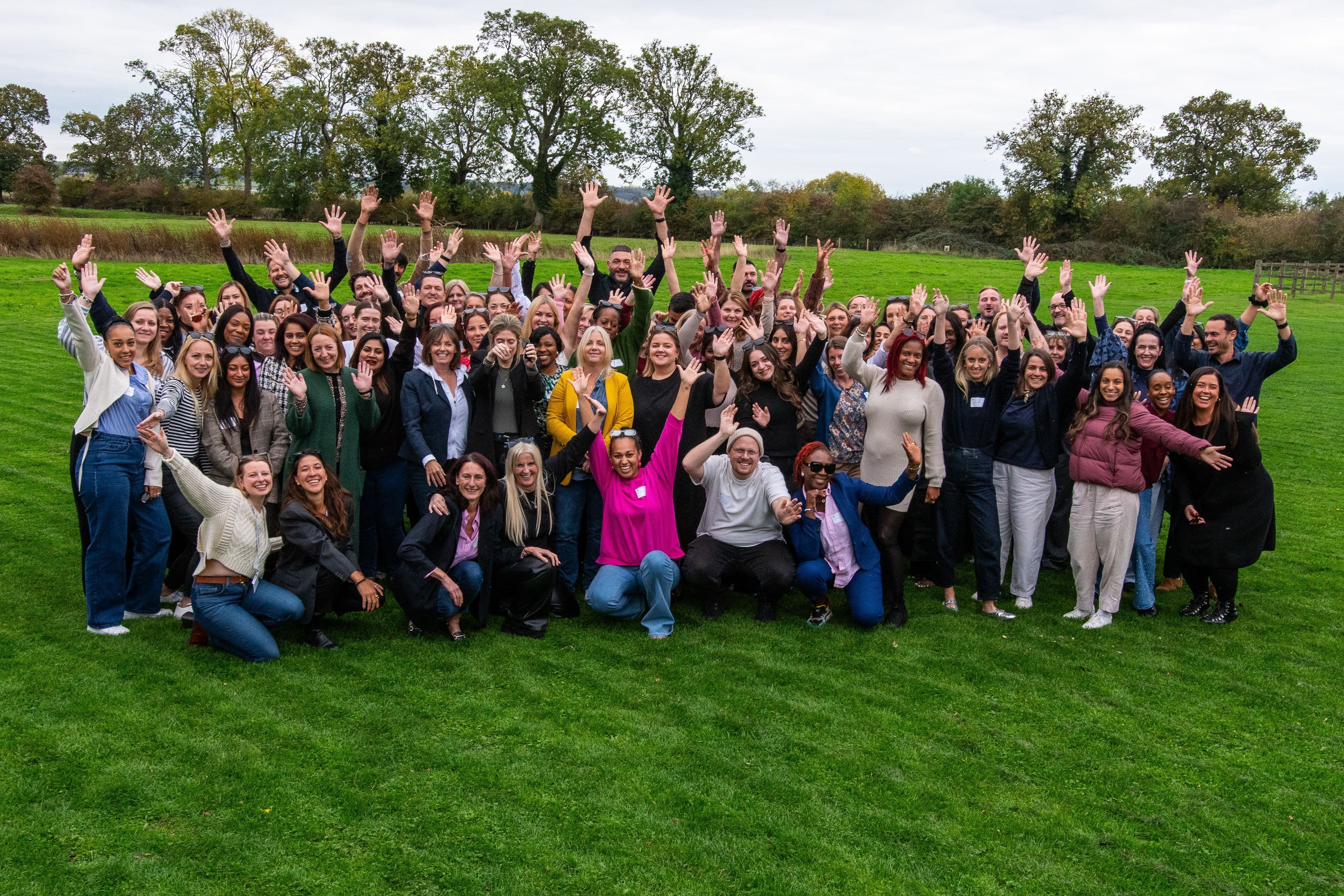 A large group of diverse people smiling and waving their hands while posing outdoors on a grassy field.