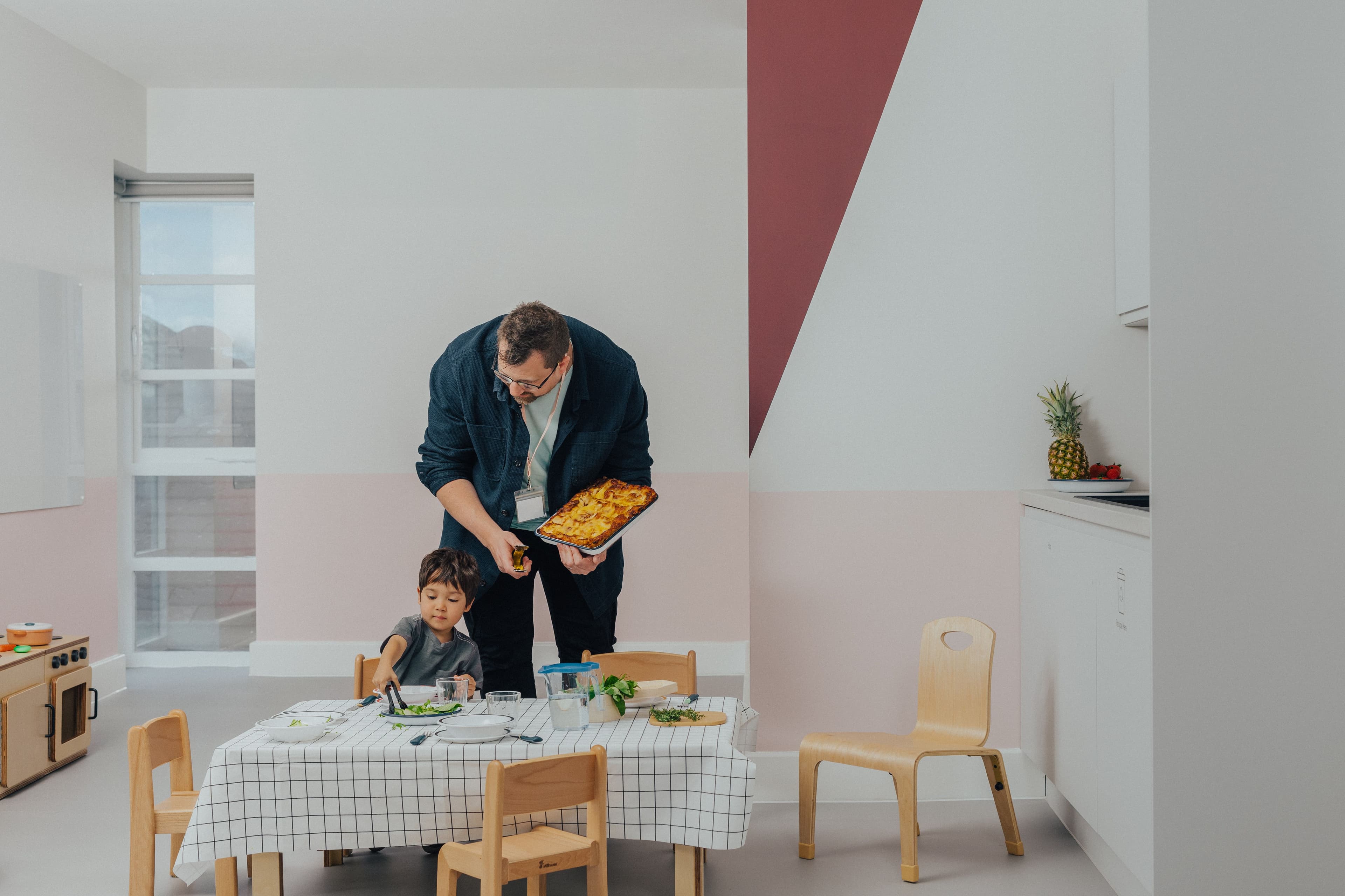 A man carries a casserole to a dining table where a child serves food from a bowl with tongs.