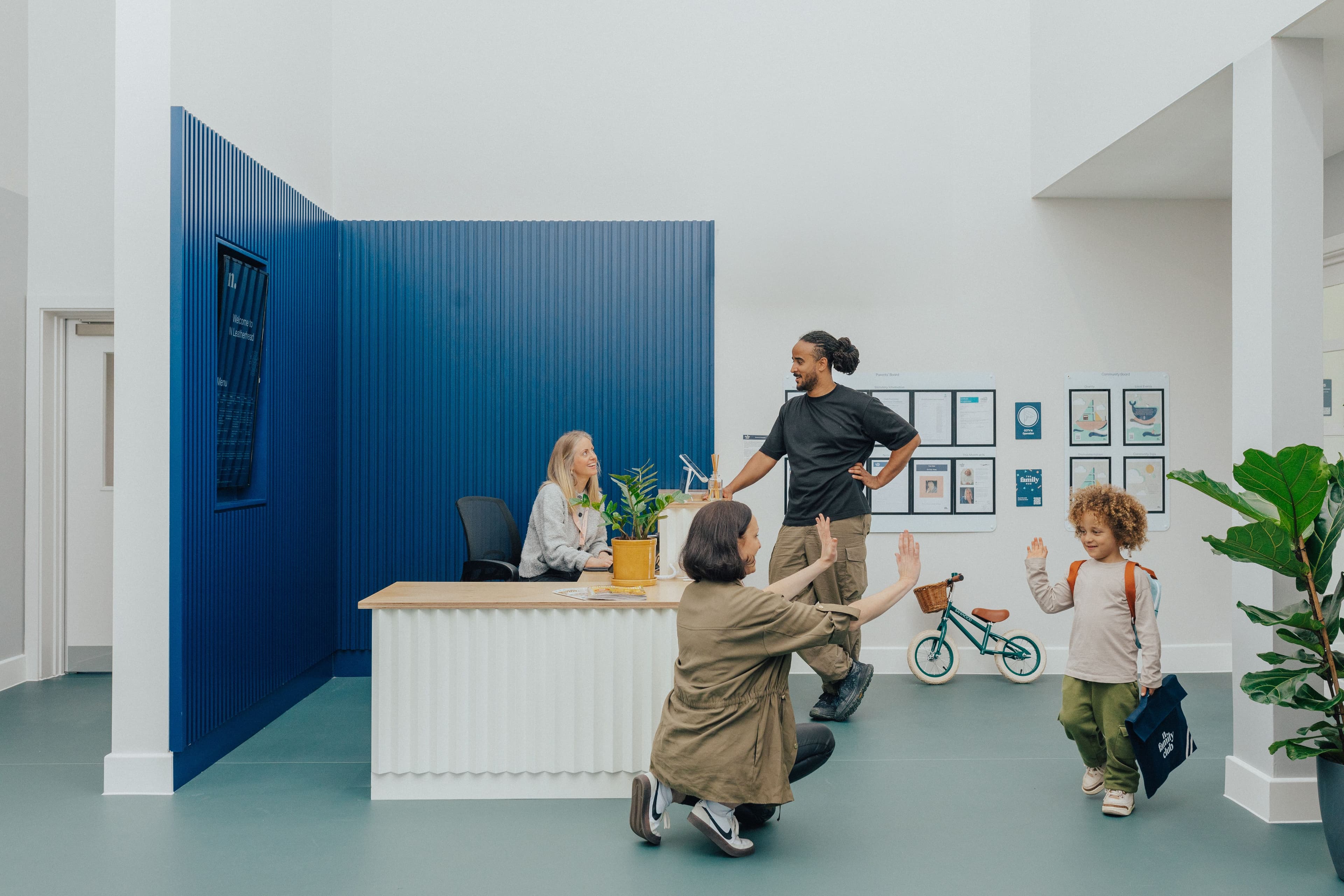 A child in a backpack high-fives a crouching adult at a modern reception desk, with another adult and staff member nearby.
