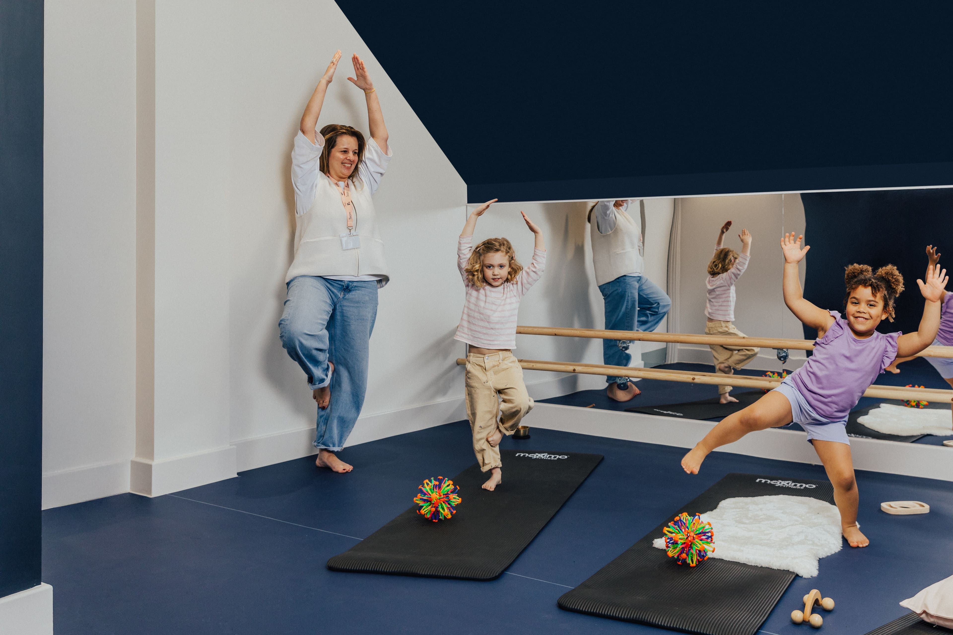 An adult and two girls happily doing balancing poses in a bright studio with a mirror and barre.