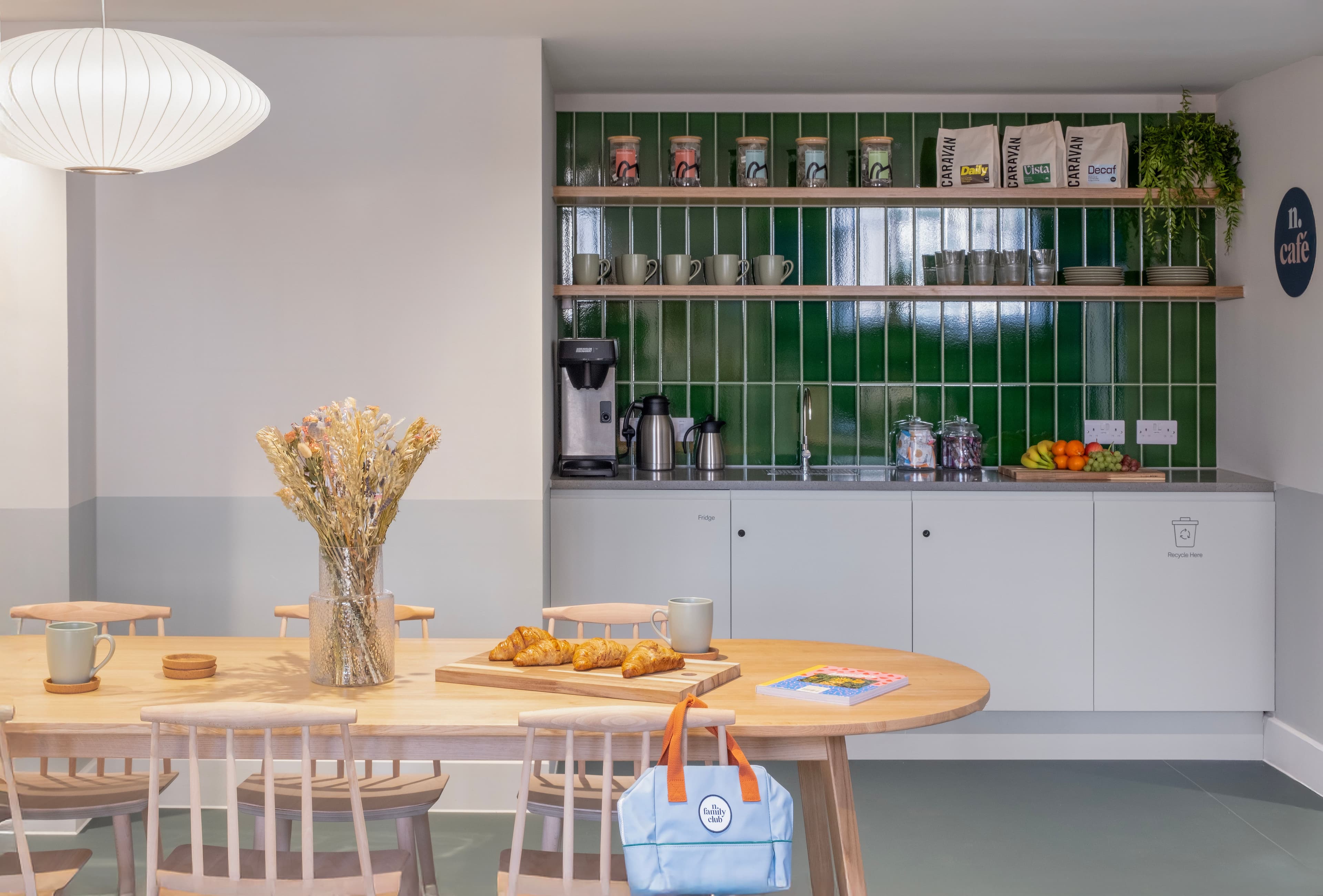 A bright cafe area with a long wooden table set with croissants and dried flowers, facing a green-tiled kitchenette with coffee supplies and fruit.