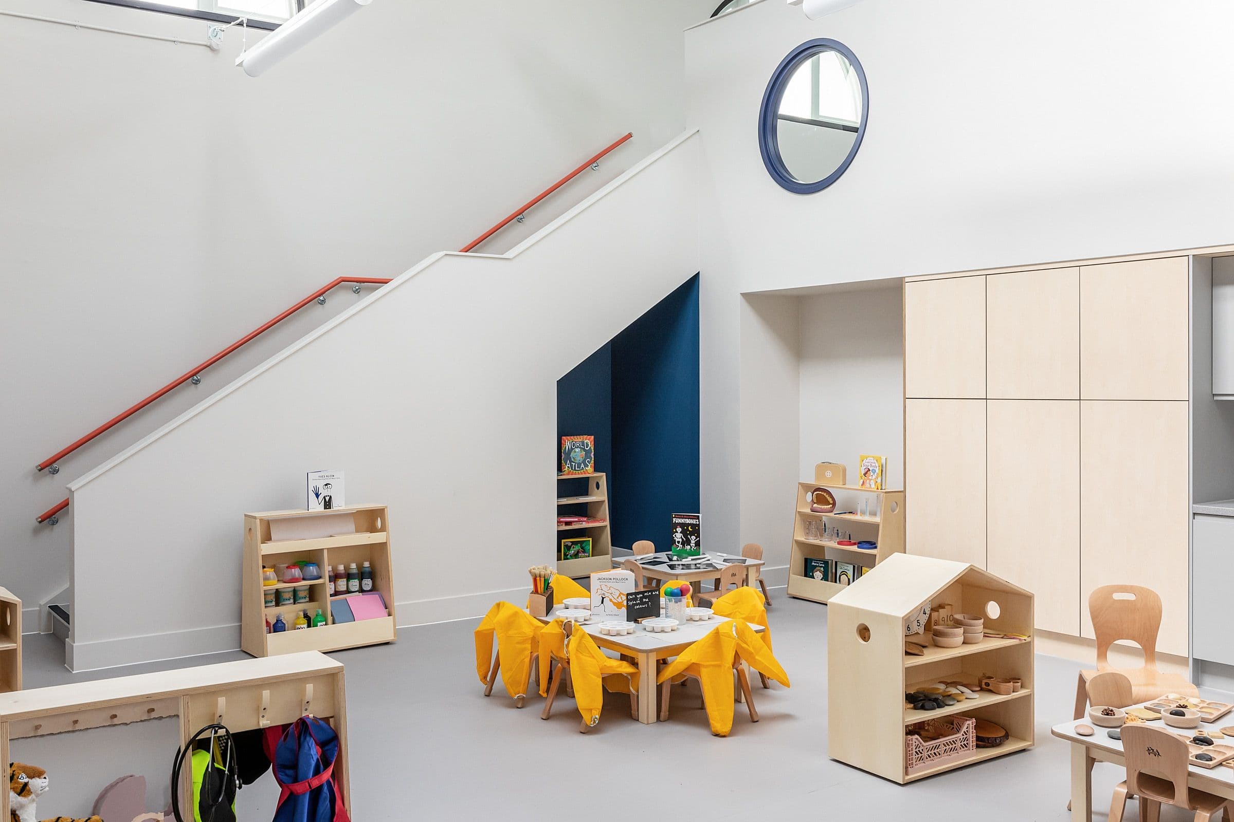 A bright, modern preschool room featuring light wood furniture, small tables with yellow smocks, a white staircase with red handrails, and a blue accent wall.