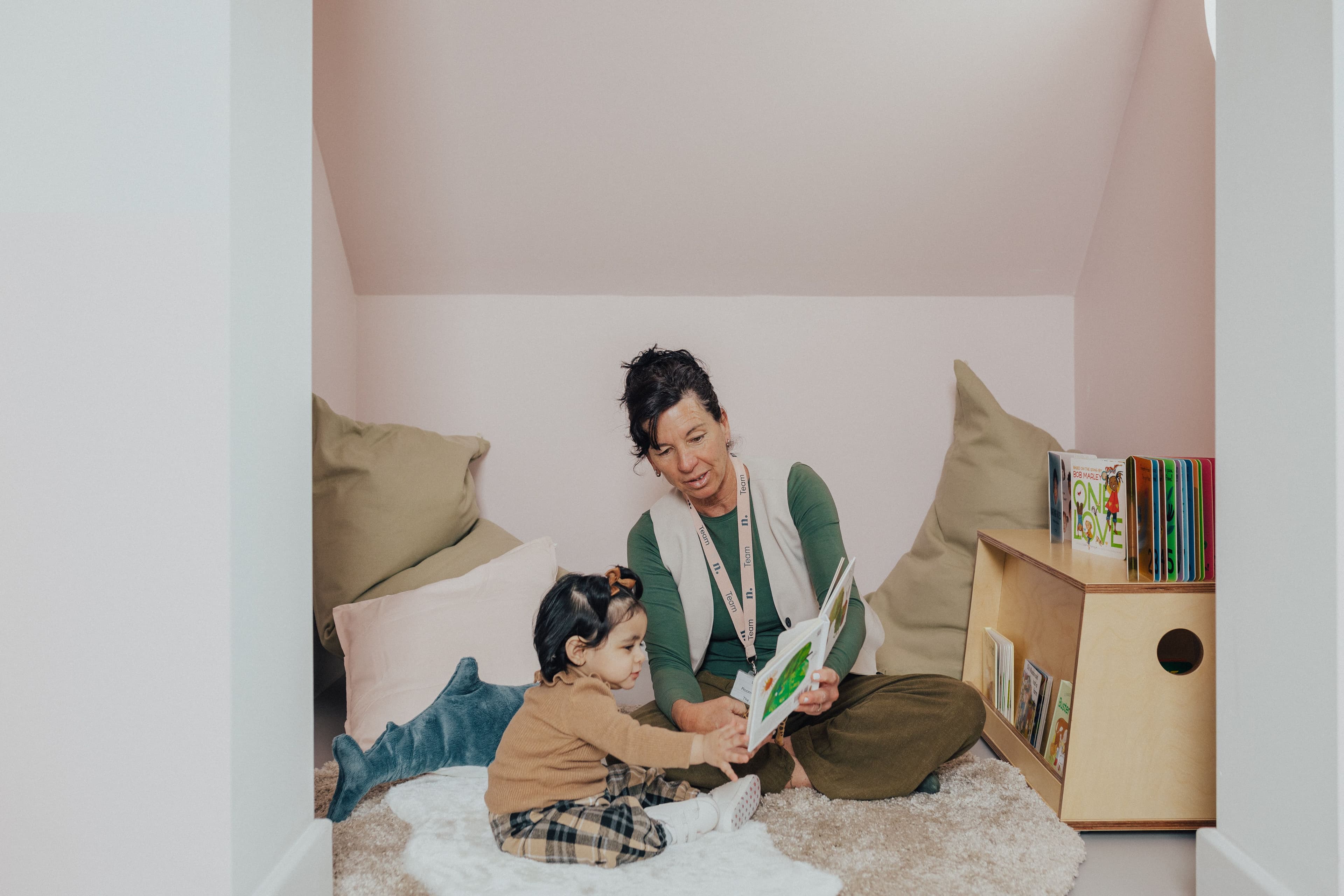 An adult reads a book to a young child in a cozy pink nook.